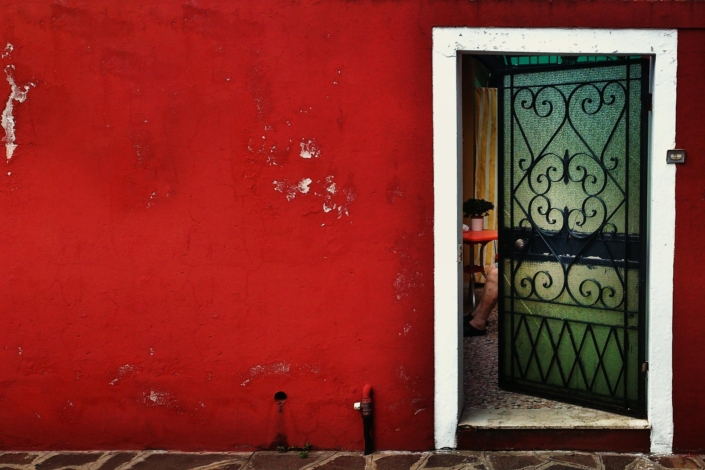 Casa rossa di Burano con porta semi-aperta da cui si vedono due gambe di anziana